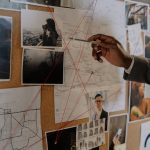 Detective examines a corkboard with maps and photos to solve a mystery.